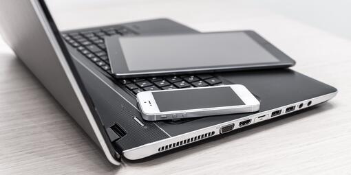 Ein Laptop steht aufgeklappt auf einem Tisch. Darauf liegt ein Tablet und daneben ein Handy.