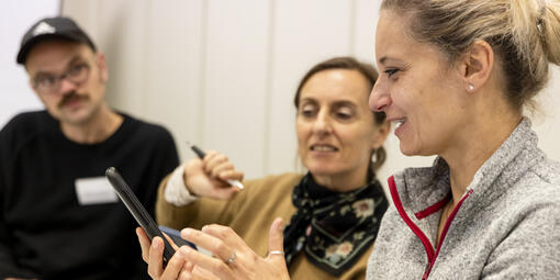 Zwei Frauen schauen gemeinsam auf ein Smartphone. Sie haben einen fröhlichen Gesichtsausdruck. Dahinter steht ein junger Mann.