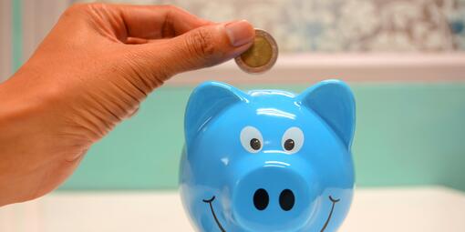 Ein blaues Sparschwein mit dem Gesicht nach vorn. Es grinst. Darüber steckt eine Hand eine Münze rein.