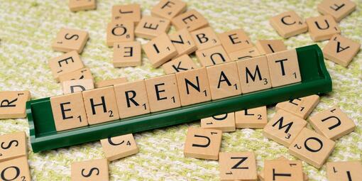 Verschiedene kleine Holzquadrate mit Buchstaben darauf. In der Mitte, innerhalb der vielen Quadrate, ist eine grüne, schmale Leiste aufgestellt. Sie zeigt das Wort "Ehrenamt".