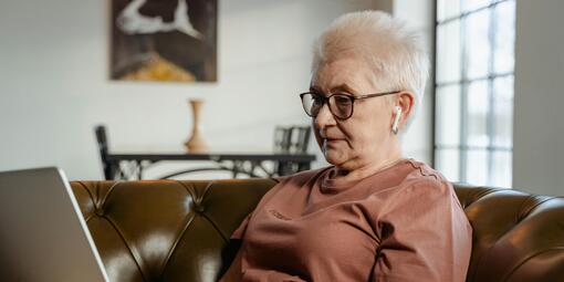 Eine ältere Dame sitzt auf dem Sofa. Sie trägt Kopfhörer und nutzt ihren Laptop.
