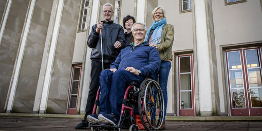 Vier personen, drei Stehend, eine sizend in einem Rollstühl stehen auf einem Patz 