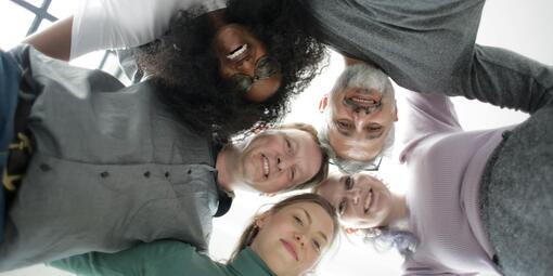 Männer und Frauen stehen Arm in Arm im Kreis. Die jeweiligen Gesichter sind von unten fotografiert.