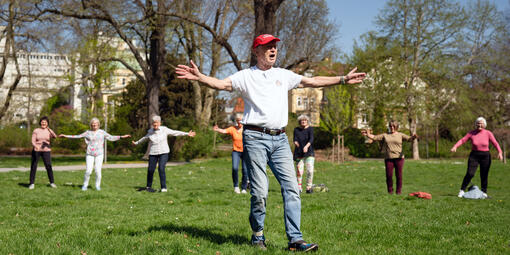 Ein Mann macht im Grünen verschiedene Sportübungen vor. Er streckt die Arme zur Seite. Einige Personen stehen dahinter und machen es nach.
