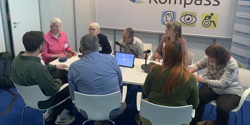 Sitzgruppe am Messestand. Einige Personen sitzen verteilt am Tisch. Es findet ein Vortrag zum iPhone statt. 