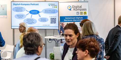 Messestand des Digital-Kompass. Im Hintergrund ein Bildschirm sowie ein Aufsteller des Projektes. Davor viele Personen im Austausch. 