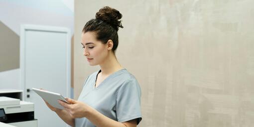 Eine junge Arzthelferin im Kasak steht hinter einem Empfangsthresen und bedient ein Tablet. 