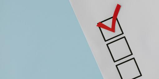 Ein weißes Blatt ist zur Hälfte zu sehen. Es liegt auf einem hellblauen Hintergrund. Auf dem Blatt sind drei Kästchen. Eines davon ist abgehakt. 