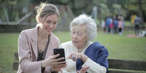 Eine junge Frau und eine ältere Dame sitzen auf einer Parkbank. Die jüngere Frau hält ein Smartphone in der Hand und zeigt der älteren Dame grinsend etwas. Die ältere Dame sieht interessiert aus und zeigt auf das Smartphone.