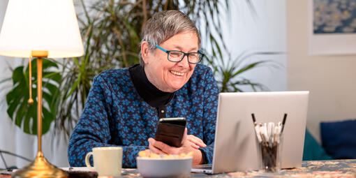 Eine Frau sitzt vor dem Laptop und hat ein Smartphone in der Hand.