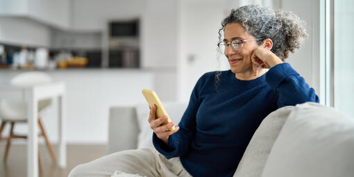Eine ältere Dame sitzt fröhlich auf dem Sofa und schaut auf ihr Handy.