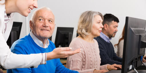 Ein Computerkabinett, in dem mehrere Personen sitzen. Im Vordergrund ist ein älterer Herr der zu einer Dame schaut, die ihm etwas am PC erklärt.