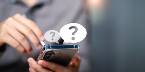 Ein Smartphone wird in der Hand gehalten. Über dem Smartphone sind zwei Sprechblasen mit jeweils einem Fragezeichen eingeblendet.