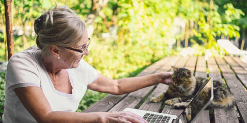 Éine ältere Dame sitzt draußen an einem Holztisch. Sieht nutzt einen Laptop und streichelt dabei eine Katze, die auf dem Tisch liegt.