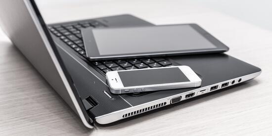 Ein Laptop steht aufgeklappt auf einem Tisch. Darauf liegt ein Tablet und daneben ein Handy.