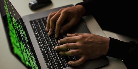 Auf einem weißen Tisch steht ein Laptop. Dieser ist aufgeklappt. Viele verschlüsselte Zeichen in grün sind darauf zu sehen. Zwei Hände tippen auf der Tastatur.