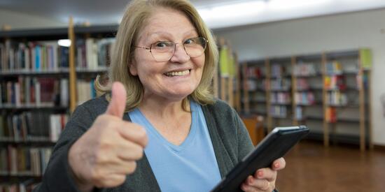 Eine ältere Dame hält ein Tablet in der Hand und zeigt einen Daumen nach oben. Sie steht vermutlich in einer Bibliothek und hat ein breites Grinsen. 