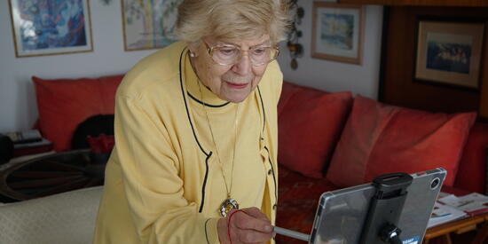 Eine ältere Dame steht vor einem Tablet, was sich in einer Halterung befindet. Sie tippt mit einem Stift darauf. 