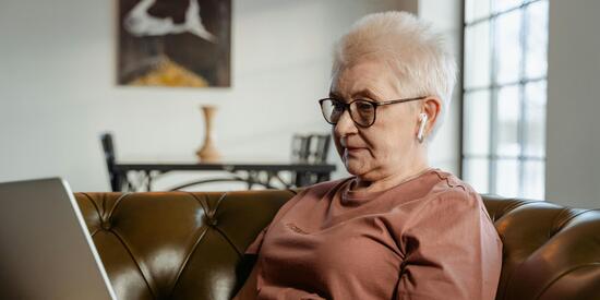 Eine ältere Dame sitzt auf dem Sofa. Sie trägt Kopfhörer und nutzt ihren Laptop.