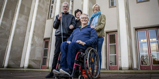 Vier personen, drei Stehend, eine sizend in einem Rollstühl stehen auf einem Patz 