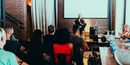 Personen sitzen auf Stühlen und sind von hinten zu sehen. Vor ihnen ist eine Bühne, auf der ein Mann auf einem Stuhl sitzt. Er hält ein Mikro in der Hand und spricht zum Publikum. 