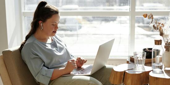 Eine junge Frau mit Down-Syndrom sitzt auf dem Sofa. Sie trägt Kopfhörer und nutzt einen Laptop auf ihrem Schoß. 