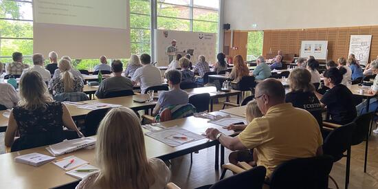 Im Konferenzsaal sitzen Personen an den Tischen. Sie sind von hinten zu sehen. Vorn eine große Leinwand mit Referent. 