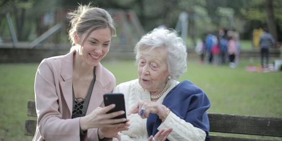 Eine junge Frau und eine ältere Dame sitzen auf einer Parkbank. Die jüngere Frau hält ein Smartphone in der Hand und zeigt der älteren Dame grinsend etwas. Die ältere Dame sieht interessiert aus und zeigt auf das Smartphone.