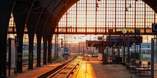 Das Bild zeigt einen Bahnhof mit einfahrendem Zug bei Sonnenaufgang