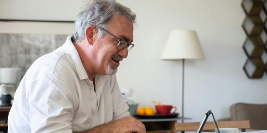Ein älterer Mann sitzt fröhlich am Schreibtisch. Vor ihm ist ein Tablet aufgestellt, auf das er scheint.