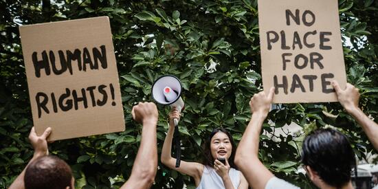 Eine Frau hält ein Megafon in die Luft und spricht in ein Aufnahmegerät. Vor ihr stehen zwei Männer, ihr Rücken ist zu sehen. Sie halten Schilder hoch. Eines mit der Schrift "Humans Rights", das andere mit "No place for hate".