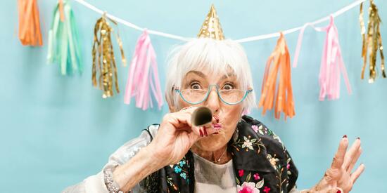 Eine ältere Frau hat einen Partyhut auf und eine Papiertröte im Mund. Sie hat die Augen weit geöffnet und ist leicht nach vorne gelehnt. Vor dem blauen Hintergrund hängt eine Girlande mit vielen bunten Elementen.