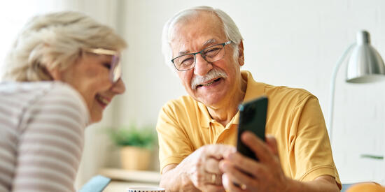 Zwei ältere Personen sitzen am Tisch. Der ältere Herr zeigt der älteren Dame etwas auf dem Handy und beide schauen fröhlich.