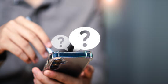 Ein Smartphone wird in der Hand gehalten. Über dem Smartphone sind zwei Sprechblasen mit jeweils einem Fragezeichen eingeblendet.