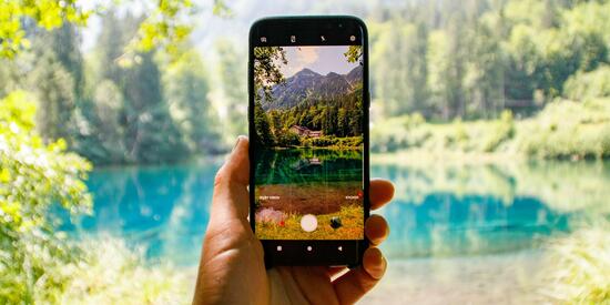 Ein Handy fotografiert einen blauen See im Wald. Im Vordergrund das Smartphone mit der Aufnahme, im Hintergrund die besagte Szene.